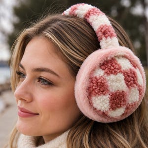 May include: Fuzzy earmuffs featuring a checkered pattern in dusty rose pink and cream white. The headband and circular ear covers are constructed from a soft, textured material, with a solid pink border surrounding the checkered center panels.