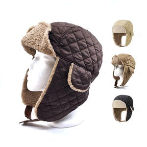 May include: A brown quilted trapper hat with a faux fur lining and ear flaps. The hat is displayed on a white mannequin head. Additional hats in beige, brown, and black are shown in the background. The hat has a leather strap.