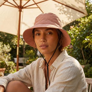 May include: A dusty rose-colored bucket hat with a wide brim and a black chin strap. The hat is made of a soft, textured material. The hat is designed for sun protection and outdoor activities.