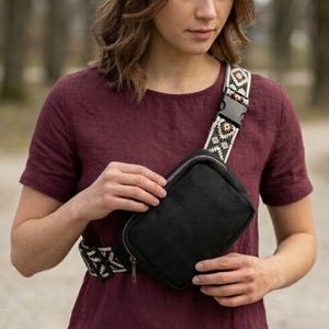 May include: Black crossbody bag with a patterned strap. The bag has a rectangular shape with a zipper closure. The strap features a geometric design in shades of white, green, and brown. The person in the image is wearing a maroon shirt.