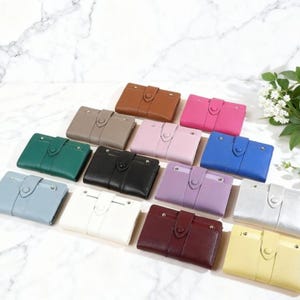 May include: A collection of leather wallets in various colors, including brown, pink, blue, and green. Each wallet has a rectangular shape with a snap closure and a small strap detail. The wallets are arranged on a white marble surface.