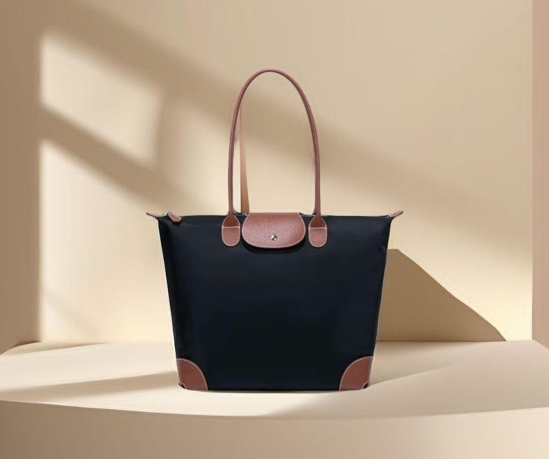 May include: A black tote bag with brown leather accents. The bag has a rectangular shape with a flap closure and two long handles. The corners of the bag are also reinforced with brown leather.