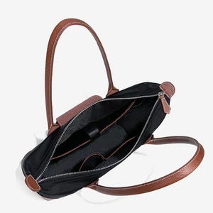 May include: Black tote bag with brown leather handles and accents. The bag has a zippered closure and multiple interior pockets. The handles are stitched with white thread. The bag is open, revealing the interior.