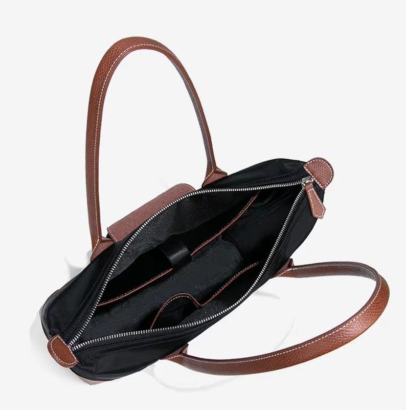 May include: Black tote bag with brown leather handles and accents. The bag has a zippered closure and multiple interior pockets. The handles are stitched with white thread. The bag is open, revealing the interior.