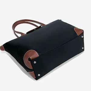 May include: Black tote bag with brown leather accents on the corners and handles. The bag has a rectangular shape with a top zipper closure. The handles are curved and attached to the top of the bag. The bag is made of a durable material.