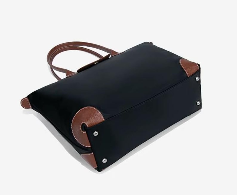 May include: Black tote bag with brown leather accents on the corners and handles. The bag has a rectangular shape with a top zipper closure. The handles are curved and attached to the top of the bag. The bag is made of a durable material.