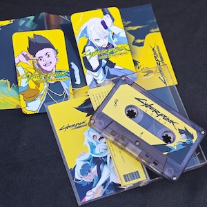 Puede incluir: Una cinta de cassette amarilla y negra con el texto "Cyberpunk 2077" impreso en ella. La cinta de cassette está rodeada de tarjetas promocionales amarillas y negras para el videojuego Cyberpunk Edgerunners.