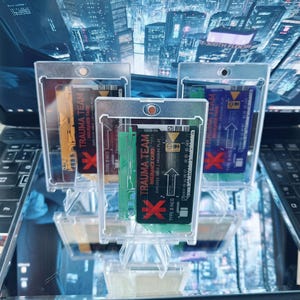 May include: Three rectangular Trauma Team insurance cards encased in clear plastic protective holders. Each card features a distinct colour scheme, including yellow, black, and blue, with printed text reading Trauma Team Insurance Card and Type 0 Neg.