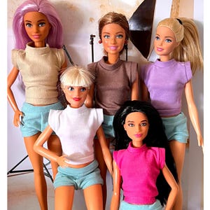 May include: Five Barbie dolls wearing different coloured tops and matching shorts. The dolls are all wearing different coloured tops, including beige, brown, purple, white, and pink. The dolls are all wearing matching shorts in a light blue colour.