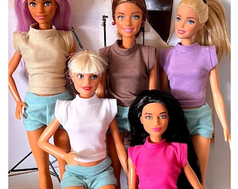 May include: Five Barbie dolls wearing different colored tops and matching shorts. The dolls are all wearing different colored tops, including beige, brown, purple, white, and pink. The dolls are all wearing matching shorts in a light blue color.