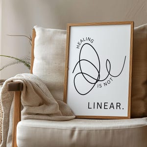 May include: A framed print with the words "HEALING IS NOT LINEAR" in black script. The artwork features a continuous line drawing in black on a white background, set in a light brown wooden frame. The print is displayed on a beige chair.
