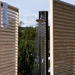 May include: A wooden outdoor shower with a slatted privacy screen. The shower head is silver and the water is running. The shower is surrounded by green plants.
