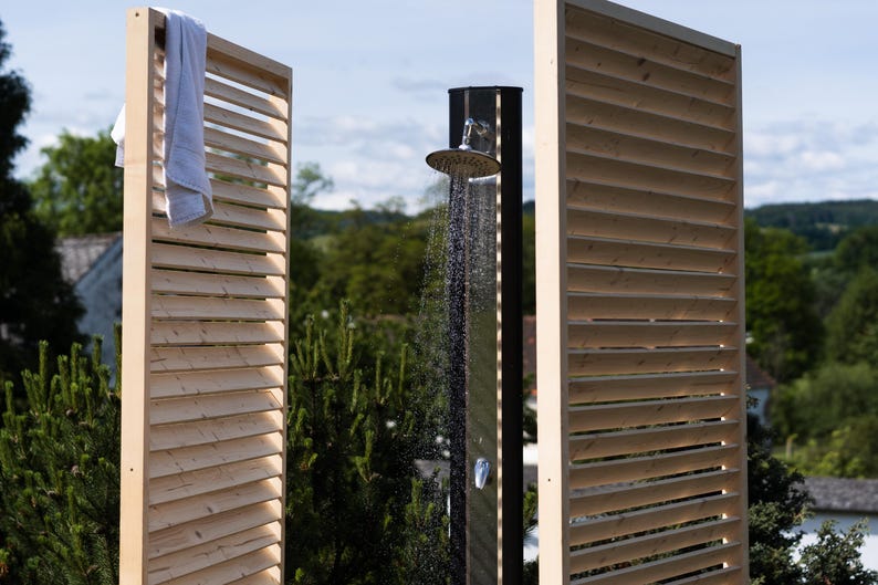 May include: A wooden outdoor shower with a slatted privacy screen. The shower head is silver and the water is running. The shower is surrounded by green plants.