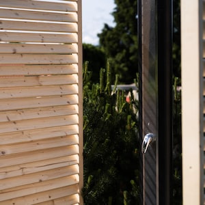 May include: A wooden slatted privacy screen with a black door. The door has a silver handle and is partially open. The screen is made of natural wood and has a light brown color.