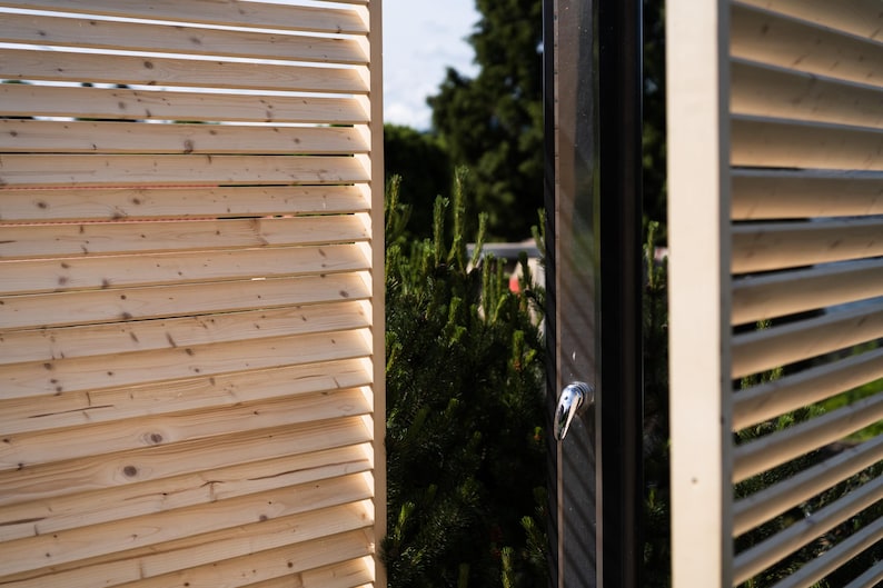 May include: A wooden slatted privacy screen with a black door. The door has a silver handle and is partially open. The screen is made of natural wood and has a light brown color.