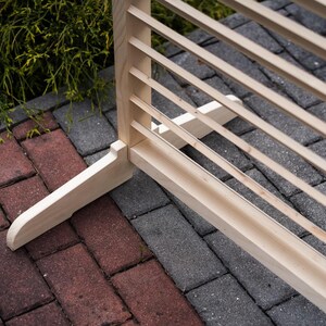 May include: A wooden slatted room divider with a natural finish. The divider has a simple design with a rectangular frame and horizontal slats. The divider is standing on a brick patio.