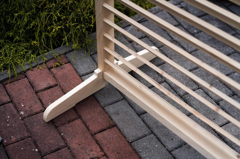 May include: A wooden slatted room divider with a natural finish. The divider has a simple design with a rectangular frame and horizontal slats. The divider is standing on a brick patio.