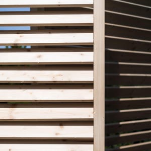 May include: Close-up of a wooden slatted fence with a light wood color on the left and a darker wood color on the right. The slats are arranged horizontally and create a pattern of light and shadow.
