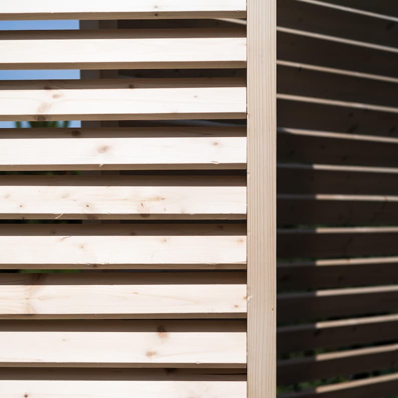 May include: Close-up of a wooden slatted fence with a light wood color on the left and a darker wood color on the right. The slats are arranged horizontally and create a pattern of light and shadow.