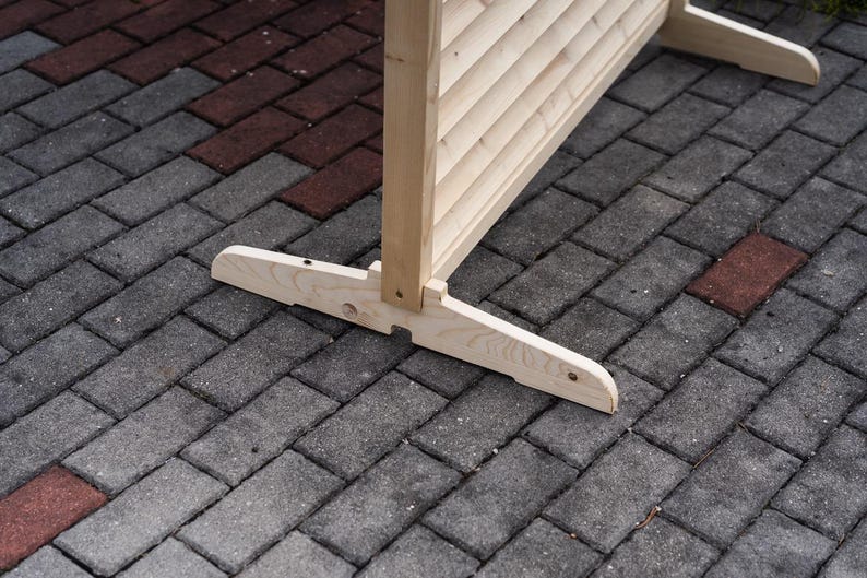 May include: A wooden room divider with a slatted design and a natural finish. The divider is standing on a gray brick patio.