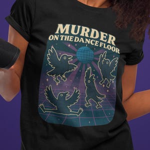 Puede incluir: Camiseta negra con el texto "MURDER ON THE DANCE FLOOR" y un gráfico de cuervos bailando. El gráfico tiene una bola de discoteca y un fondo de cuadrícula. La persona de la imagen sostiene un altavoz negro.