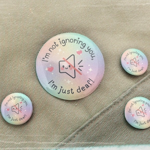 May include: Four round badges with a pastel rainbow gradient. The largest badge has a speaker icon with a line through it and the text "I'm not ignoring you, I'm just deaf!" The smaller badges repeat the same design.