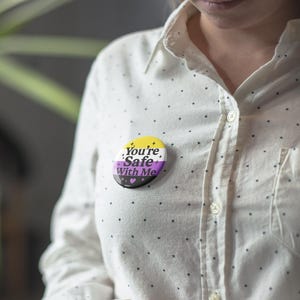 May include: A round button with the text "You're Safe With Me" in white, yellow, purple, and black. The button is pinned to a white button-down shirt with small black polka dots.