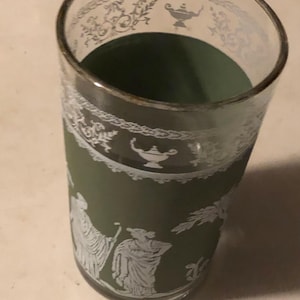 May include: A tall, clear glass with a gold rim and a green interior. The glass features white decorative patterns of figures and floral designs. The glass is likely used for drinking beverages.