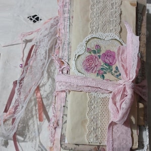 Puede incluir: Un diario de estilo vintage con una cinta rosa suave y adornos de encaje. El diario presenta un diseño floral con rosas rosas y páginas de color crema. Delicados encajes y cintas adornan la portada, creando una estética romántica y femenina. El diario está atado con una cinta rosa.