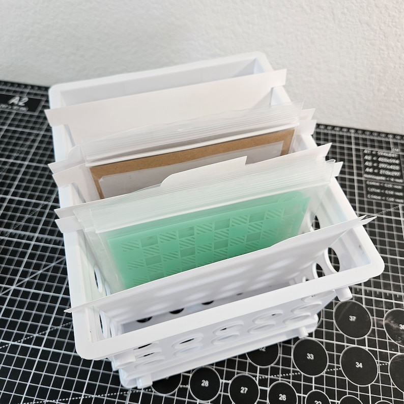 May include: A white plastic storage bin with multiple clear plastic sleeves containing various craft supplies. The sleeves are labeled with white tabs. The bin is sitting on a black and white grid surface.