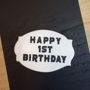 May include: White oval sign with black text that reads "Happy 1st Birthday" on a black background.