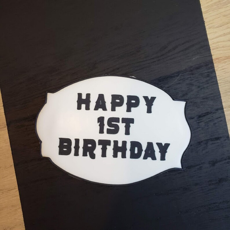 May include: White oval sign with black text that reads "Happy 1st Birthday" on a black background.