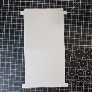 May include: A white card blank on both sides, folded in half. The card is lying flat on a black cutting mat with white grid lines.