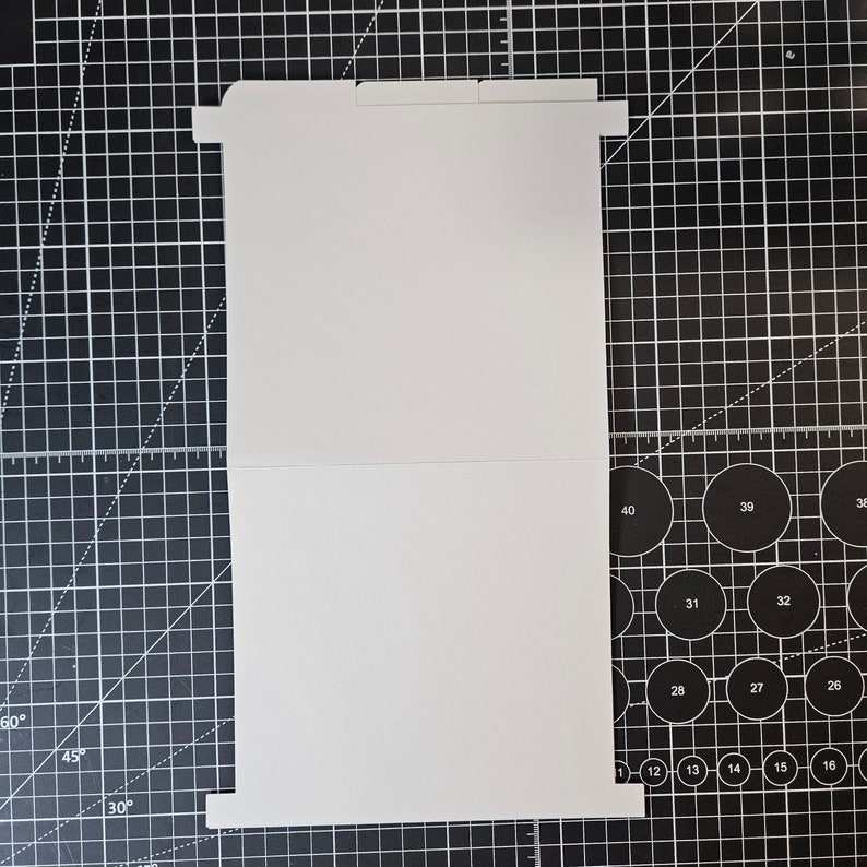 May include: A white card blank on both sides, folded in half. The card is lying flat on a black cutting mat with white grid lines.