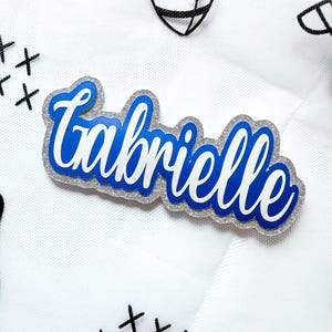 May include: A name tag with the name "Gabrielle" in a cursive font. The name is in white and blue, with a silver glitter border. The background is white with black football and cross stitch designs.