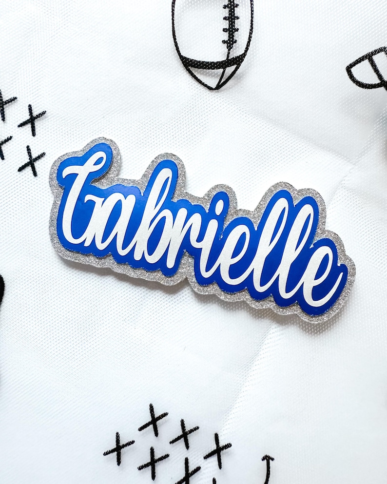 May include: A name tag with the name "Gabrielle" in a cursive font. The name is in white and blue, with a silver glitter border. The background is white with black football and cross stitch designs.