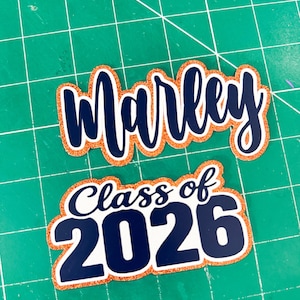 May include: Two graduation-themed cutouts. The top cutout says "Marley" in navy blue script with an orange glitter outline. The bottom cutout says "Class of 2026" in navy blue and white with an orange glitter outline.