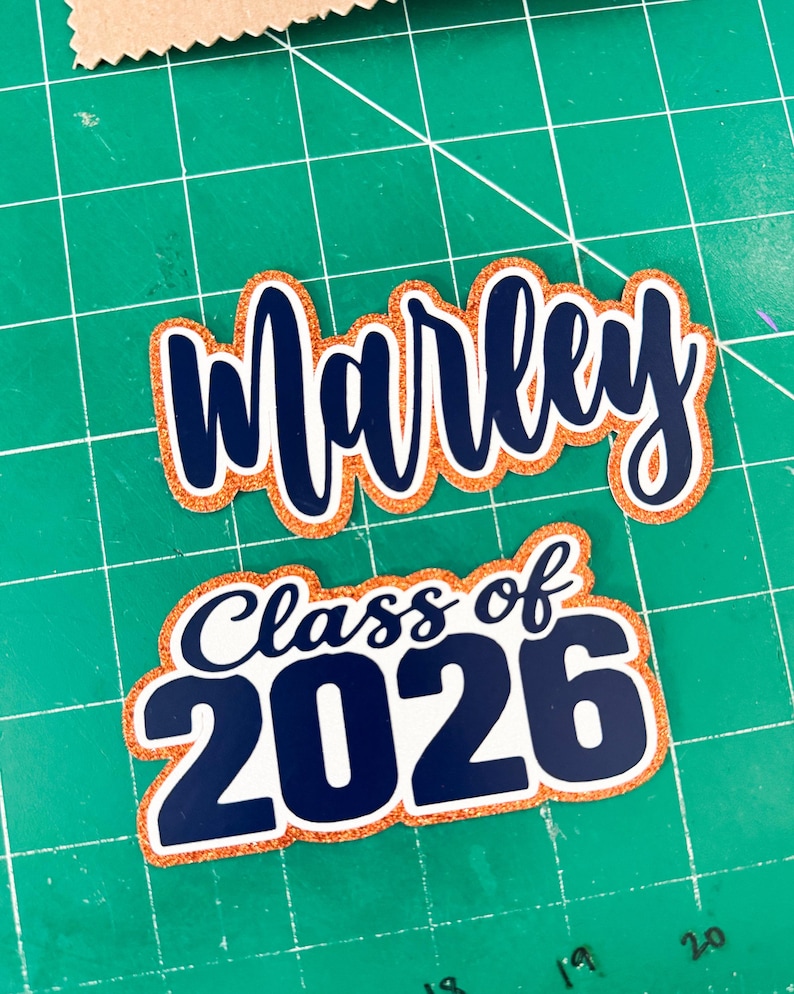 May include: Two graduation-themed cutouts. The top cutout says "Marley" in navy blue script with an orange glitter outline. The bottom cutout says "Class of 2026" in navy blue and white with an orange glitter outline.
