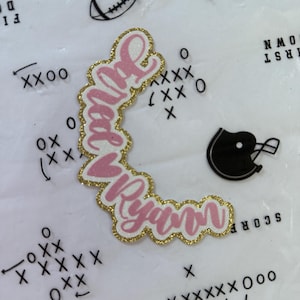 May include: A pink and gold glitter name cutout with the name "Juliet Ryan" in a cursive font. The cutout is surrounded by football-themed graphics, including a football, helmet, and play diagrams.