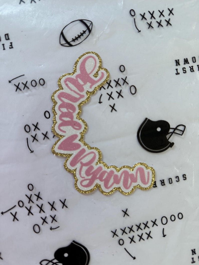 May include: A pink and gold glitter name cutout with the name "Juliet Ryan" in a cursive font. The cutout is surrounded by football-themed graphics, including a football, helmet, and play diagrams.
