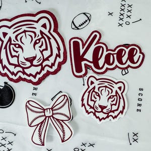 May include: A selection of maroon and white stickers with a sports theme. Includes a tiger head, the name "Kloee", a baseball bow, a football, and a football helmet. The tiger head stickers have a glittery maroon outline.