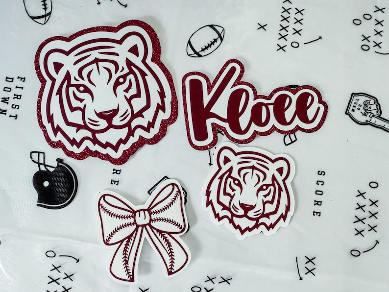 May include: A selection of maroon and white stickers with a sports theme. Includes a tiger head, the name "Kloee", a baseball bow, a football, and a football helmet. The tiger head stickers have a glittery maroon outline.