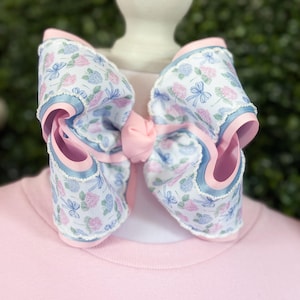 May include: A large, decorative bow with a floral pattern in pink and blue on a white background. The bow has pink and blue ribbon edging and a pink center. The bow is placed on a pink garment.