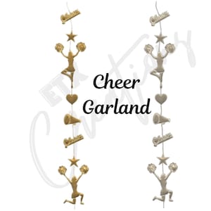 May include: Gold-colored cheer garland with cheerleader figures, stars, hearts, and megaphones. The word "Cheer" and "Garland" are written in black text. The garland is designed for decorative purposes.
