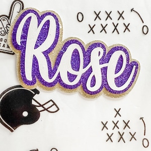 May include: A purple and gold glitter name cutout reading "Rose" in a cursive font. The name is outlined in gold glitter with a purple glitter centre. The white background features football-themed graphics.