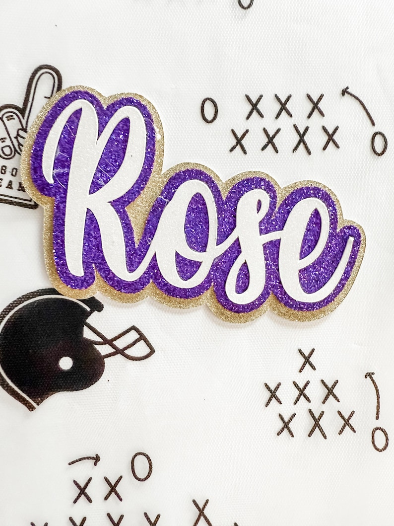 May include: A purple and gold glitter name cutout reading "Rose" in a cursive font. The name is outlined in gold glitter with a purple glitter centre. The white background features football-themed graphics.