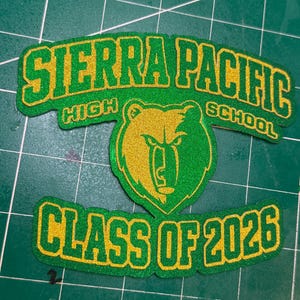 May include: A green and gold patch with the text "SIERRA PACIFIC HIGH SCHOOL" and "CLASS OF 2026". The patch features a bear graphic in the centre. The text and bear are outlined in gold.