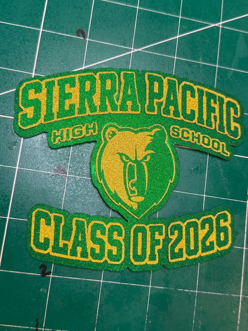May include: A green and gold patch with the text "SIERRA PACIFIC HIGH SCHOOL" and "CLASS OF 2026". The patch features a bear graphic in the centre. The text and bear are outlined in gold.