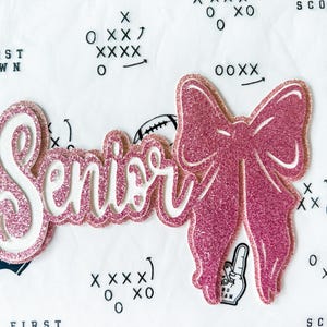 May include: A pink glitter "Senior" cutout with a matching bow. The word "Senior" is in a cursive font with a white outline. The bow is to the right of the word. The background is white with football-themed graphics.