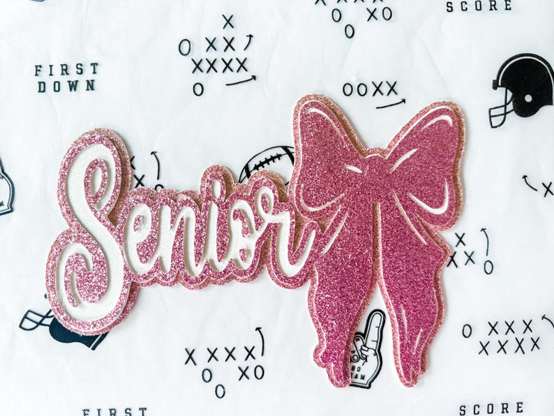 May include: A pink glitter "Senior" cutout with a matching bow. The word "Senior" is in a cursive font with a white outline. The bow is to the right of the word. The background is white with football-themed graphics.
